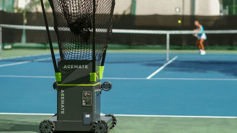 Tennis ball machine feeding a player on an outdoor hard court