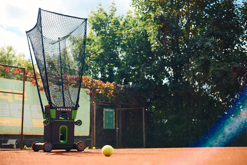 Acemate tennis ball machine set up on an outdoor clay court with a tennis ball in the foreground